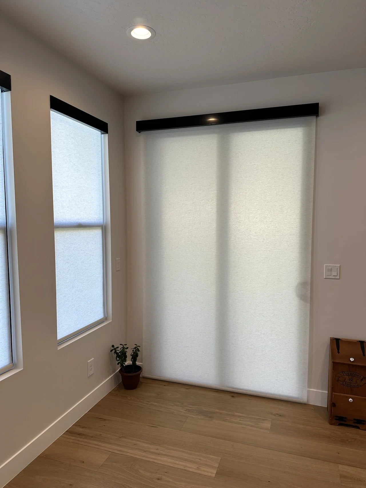 A corner of a room with three windows covered by light-colored blinds, a small potted plant on the floor near the corner, and a wooden box on the right side.