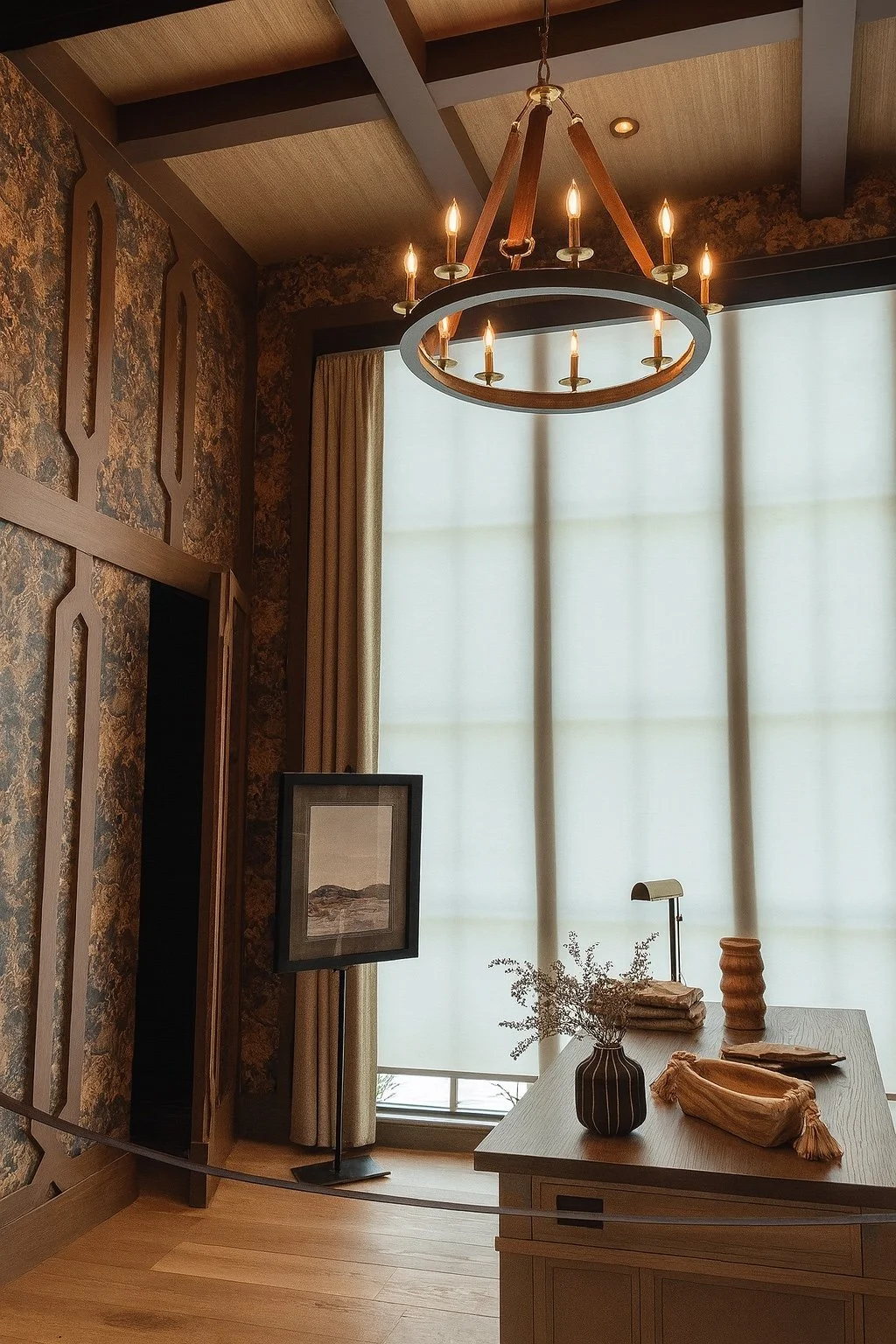 Interior of a room with a modern chandelier, wooden ceiling beams, a window with sheer curtains, a framed picture on a stand, a vase with dried flowers, and wooden decorative objects on a table.