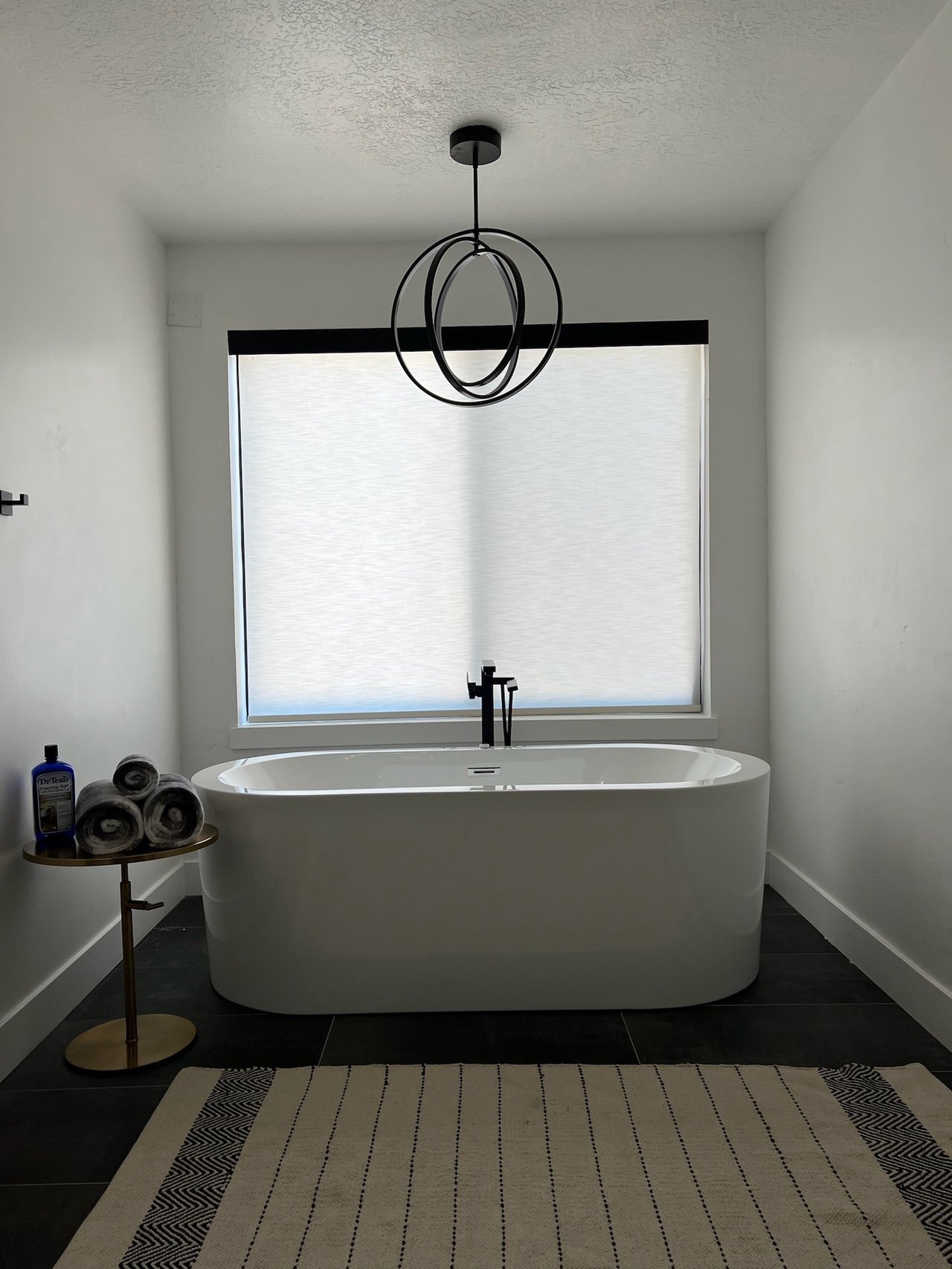 Modern bathroom with a freestanding bathtub in front of a window with shaded blinds, a small side table with rolled towels and a bottle of body wash, and a contemporary circular pendant light fixture hanging from the ceiling.