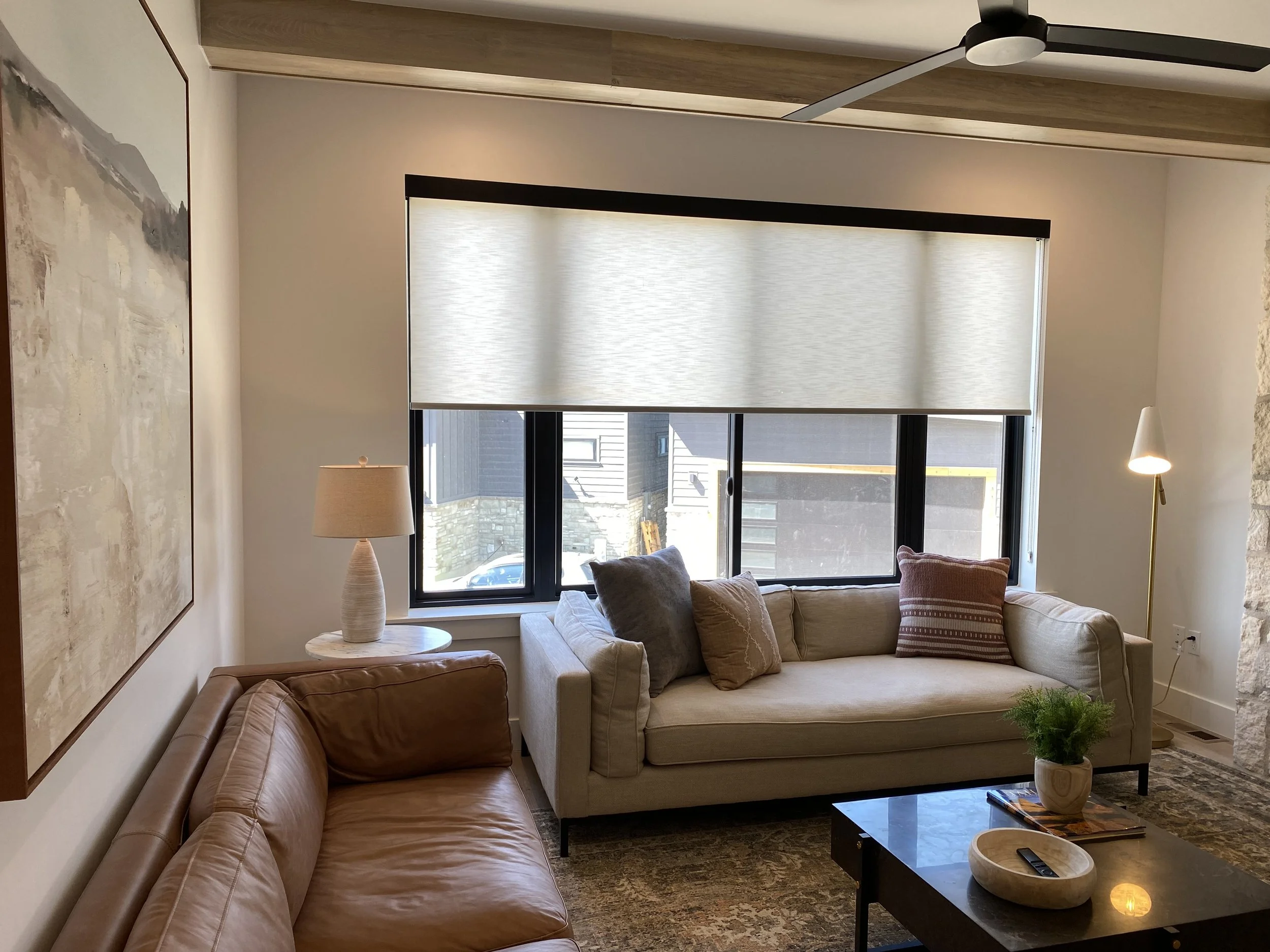Living room with two sofas, one leather and one fabric, a coffee table with a plant and remote, and large window with shades, illuminated by a standing lamp and a floor lamp, with a ceiling fan overhead.