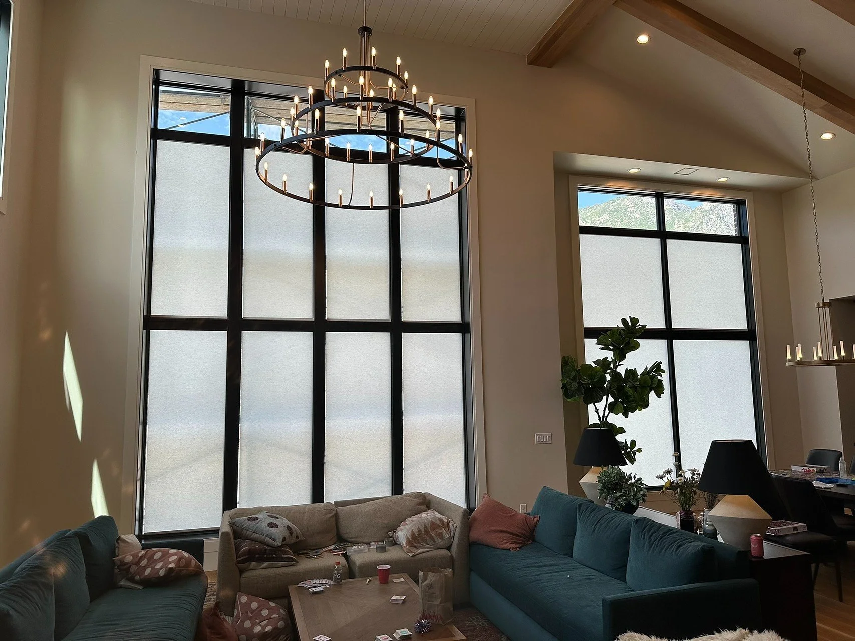 Living room with large windows, a chandelier, and multiple sofas with pillows and items on coffee table.