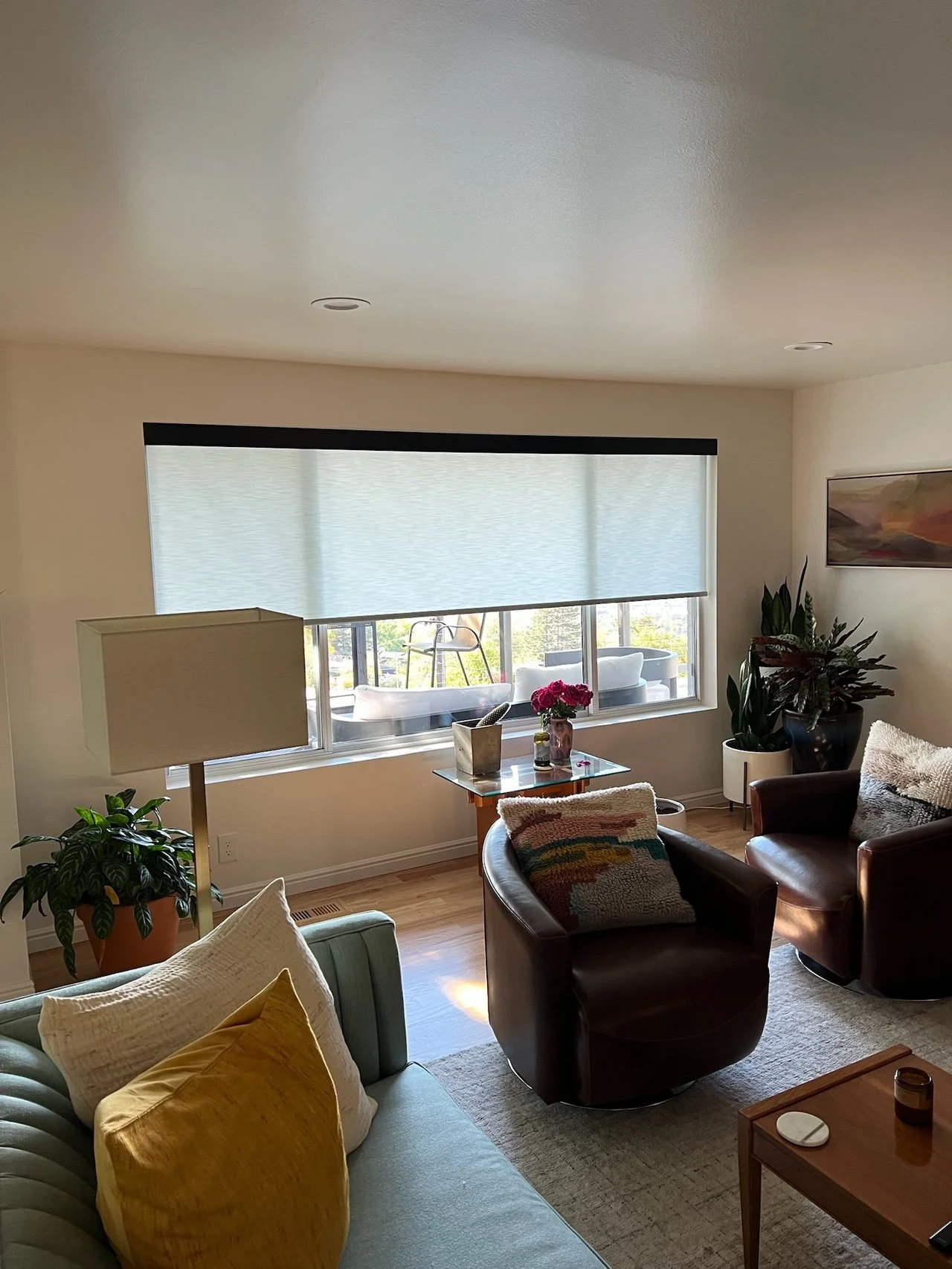Living room with large window, potted plants, a glass table with flowers, sofa, armchairs, lamps, artwork, and wooden furniture.