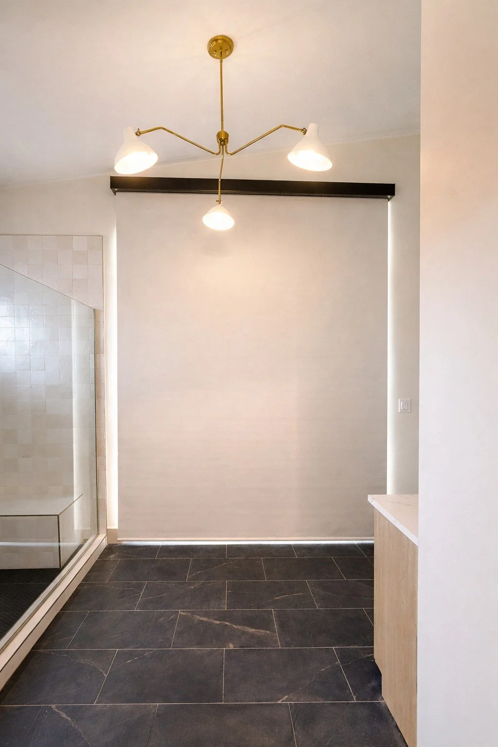 Modern bathroom with black floor tiles, a glass shower enclosure on the left, a gold ceiling light fixture with three lamps, and a white wall with a small built-in cabinet on the right.