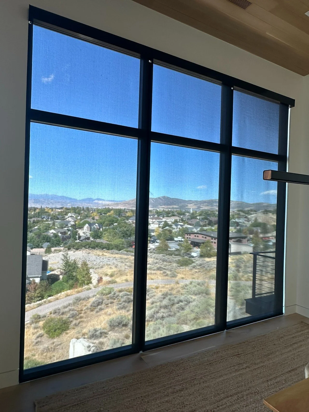 Large window with black framing showing a scenic view of houses, trees, and mountains in the distance, with a blue sky and a few clouds.
