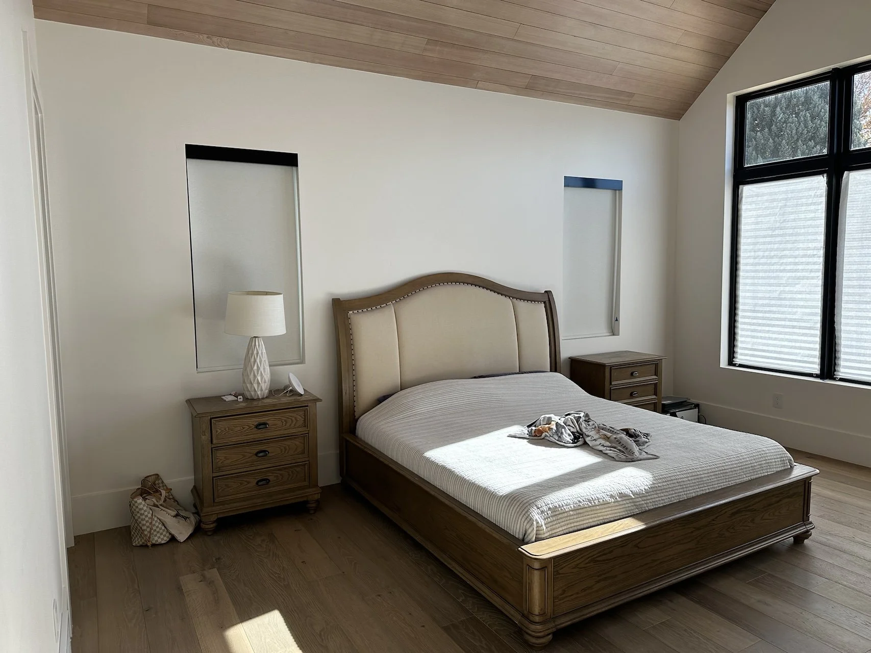 A bedroom with a wooden bed frame, bedside tables, and a lamp, with a large window and natural light.