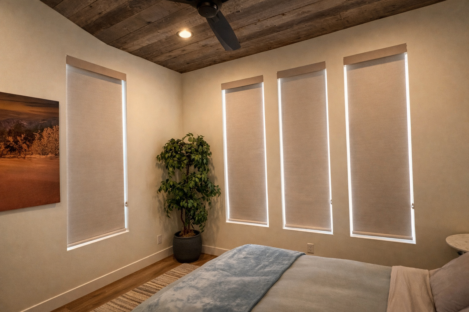 A bedroom with a wooden ceiling, four vertical windows with beige blinds, a bed with a blue blanket, a potted plant in a gray basket, a framed landscape picture, a nightstand, and a ceiling fan.