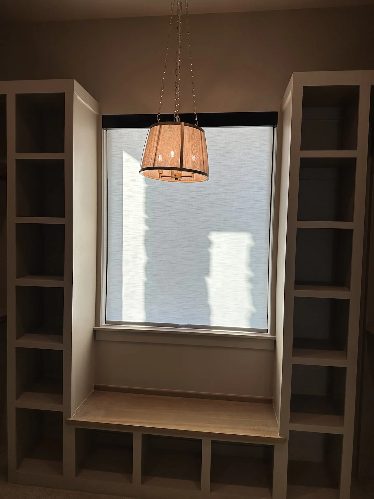 Empty built-in bookshelf with a window view, a wooden bench, and a hanging light fixture in a room.