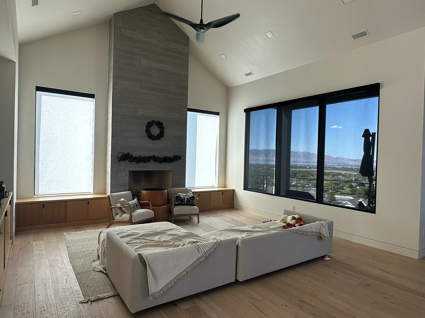 Living room with white sofa, large windows, a fireplace with a gray stone chimney, a ceiling fan, a view of mountains, and some chairs and a stuffed animal on the sofa.