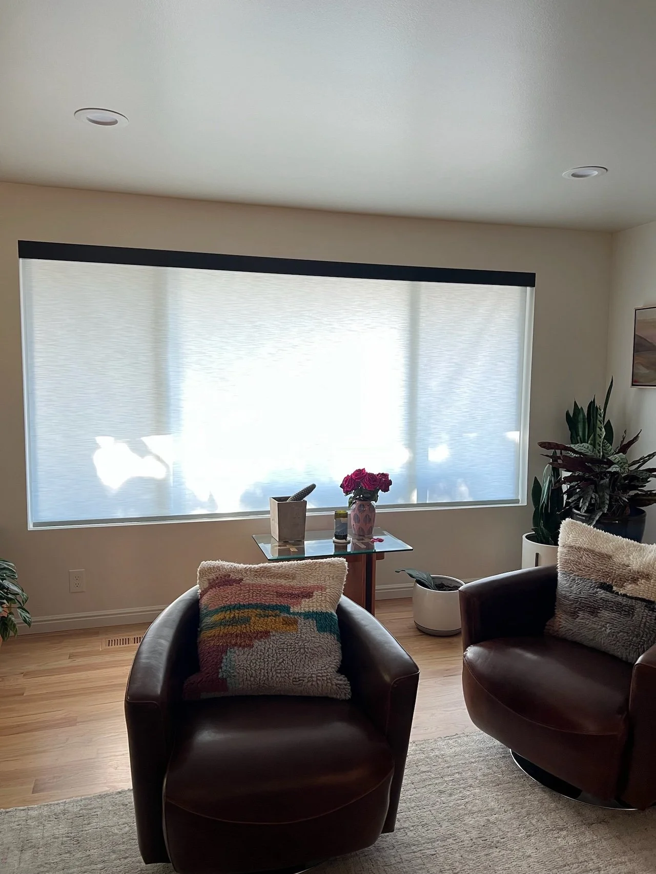 Living room with two brown leather armchairs, a small glass table with a vase of pink flowers and a few decorative items, a large window with a closed white shade, plants in pots, a painting on the wall, hardwood floor, and a beige rug.