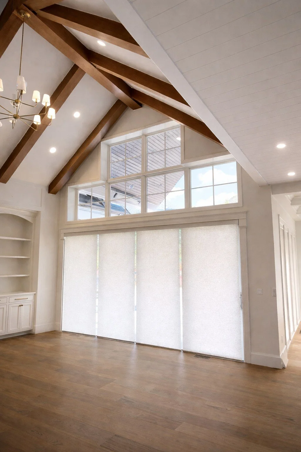 Interior of a room with vaulted ceiling, wooden beams, large windows with white blinds, hardwood flooring, and a white built-in shelf.