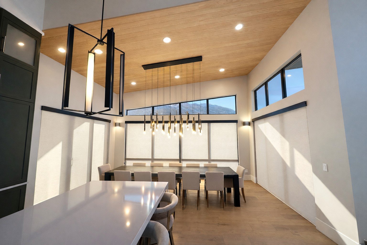 Modern dining room with wooden ceiling, large dining table, beige chairs, and unique hanging light fixtures, with large windows allowing natural light.