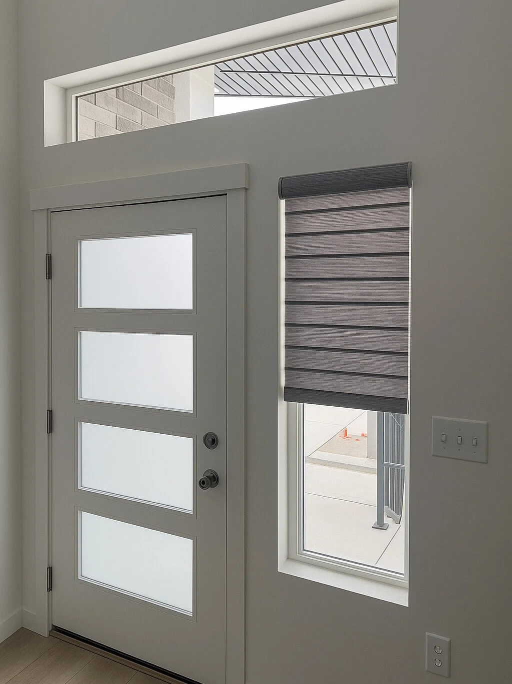 Interior view of a modern front door with four frosted glass panels, a window with a gray horizontal roller blind, an external view of a sidewalk and building, and a window above the door letting in natural light.
