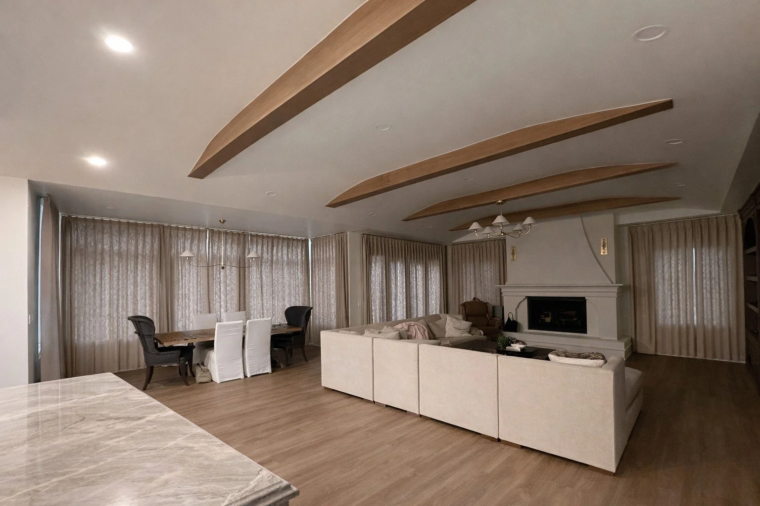 Living room with beige sectional sofa, fireplace, wooden ceiling accents, curtains, dining table with chairs, hardwood floors, and ceiling lights.