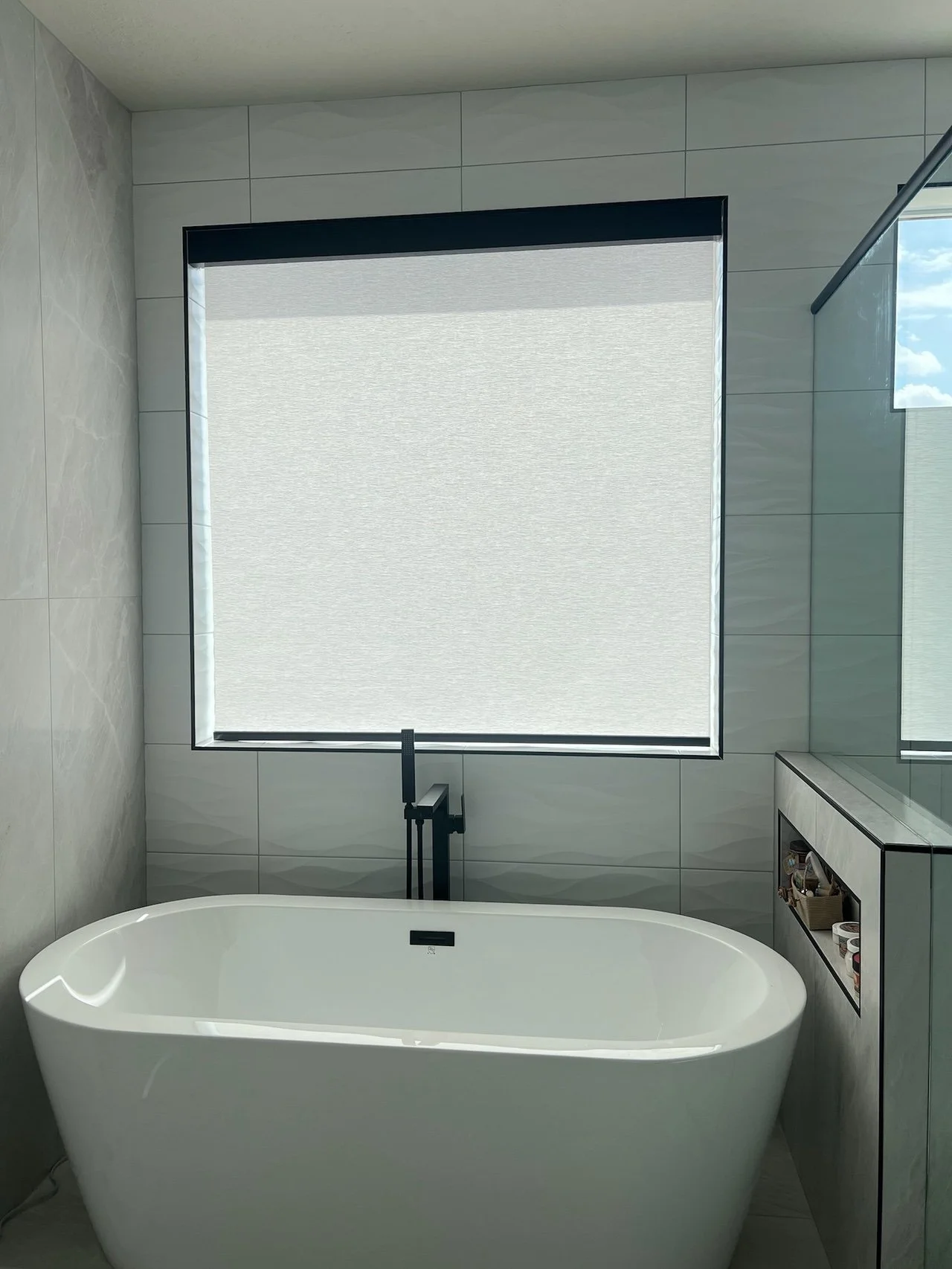 Modern bathroom with a freestanding bathtub, large window with a roller shade, gray tiled walls, and a built-in shelf with toiletries.