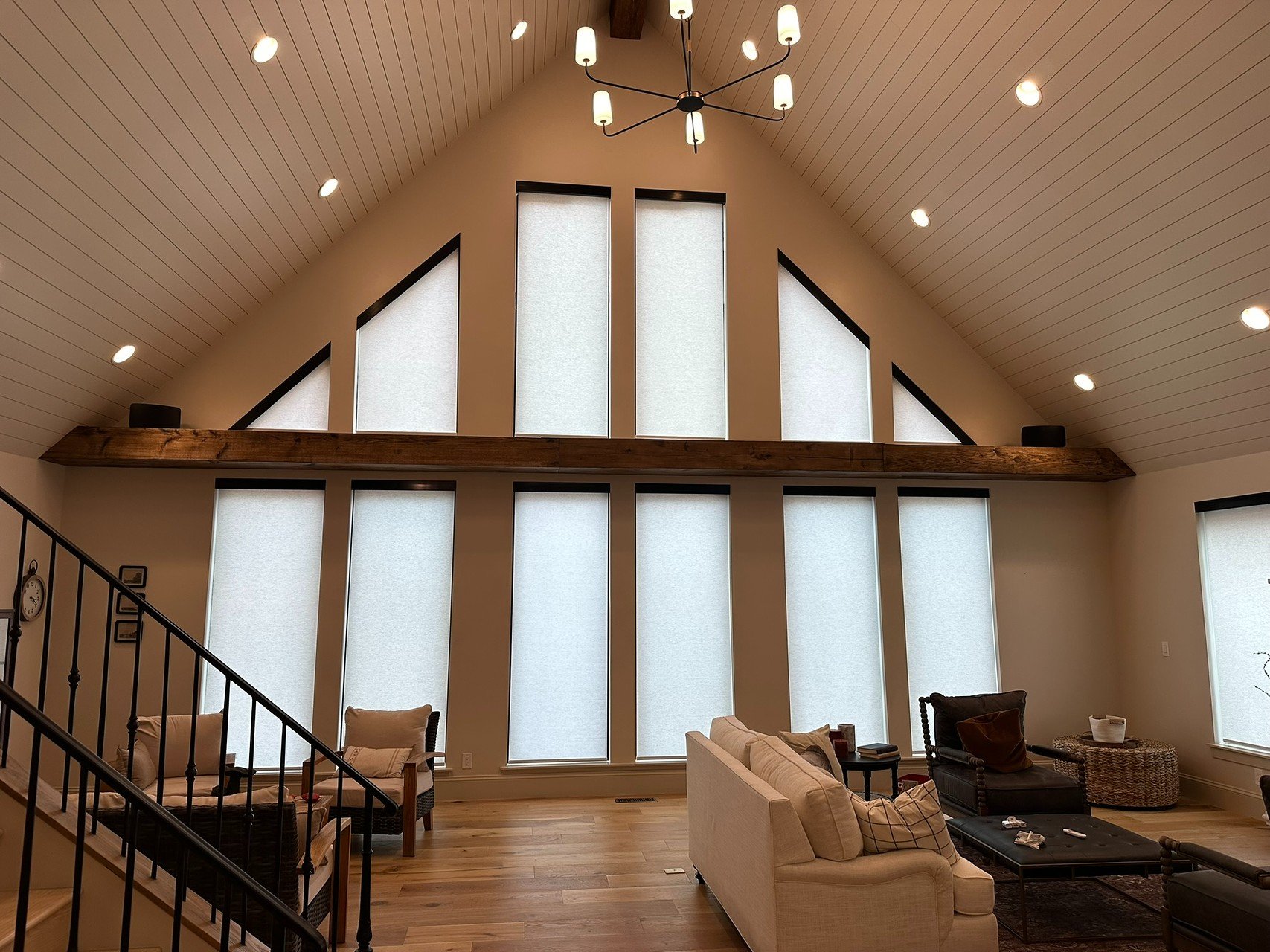Living room with high angled ceiling, large windows with white shades, and modern light fixture. Contains a beige sofa, black armchair, wooden coffee table, and other furniture on a wooden floor.