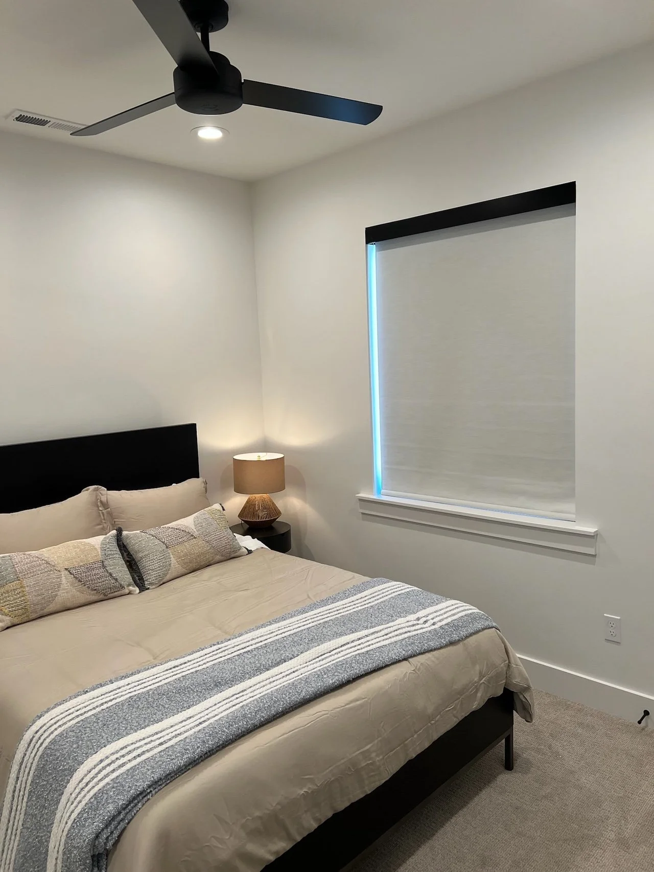 A neatly made bed with beige bedding, decorative pillows, a bedside lamp, a black ceiling fan, and a window with a closed blind in a modern bedroom.