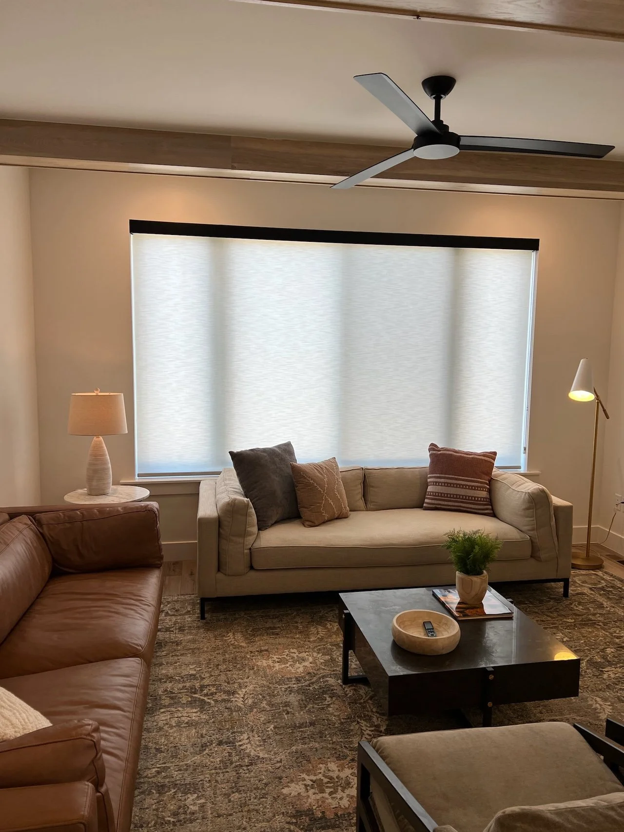 Living room with beige sofas, round coffee table, potted plant, standing lamp, table lamp, ceiling fan, and large window with light curtains.