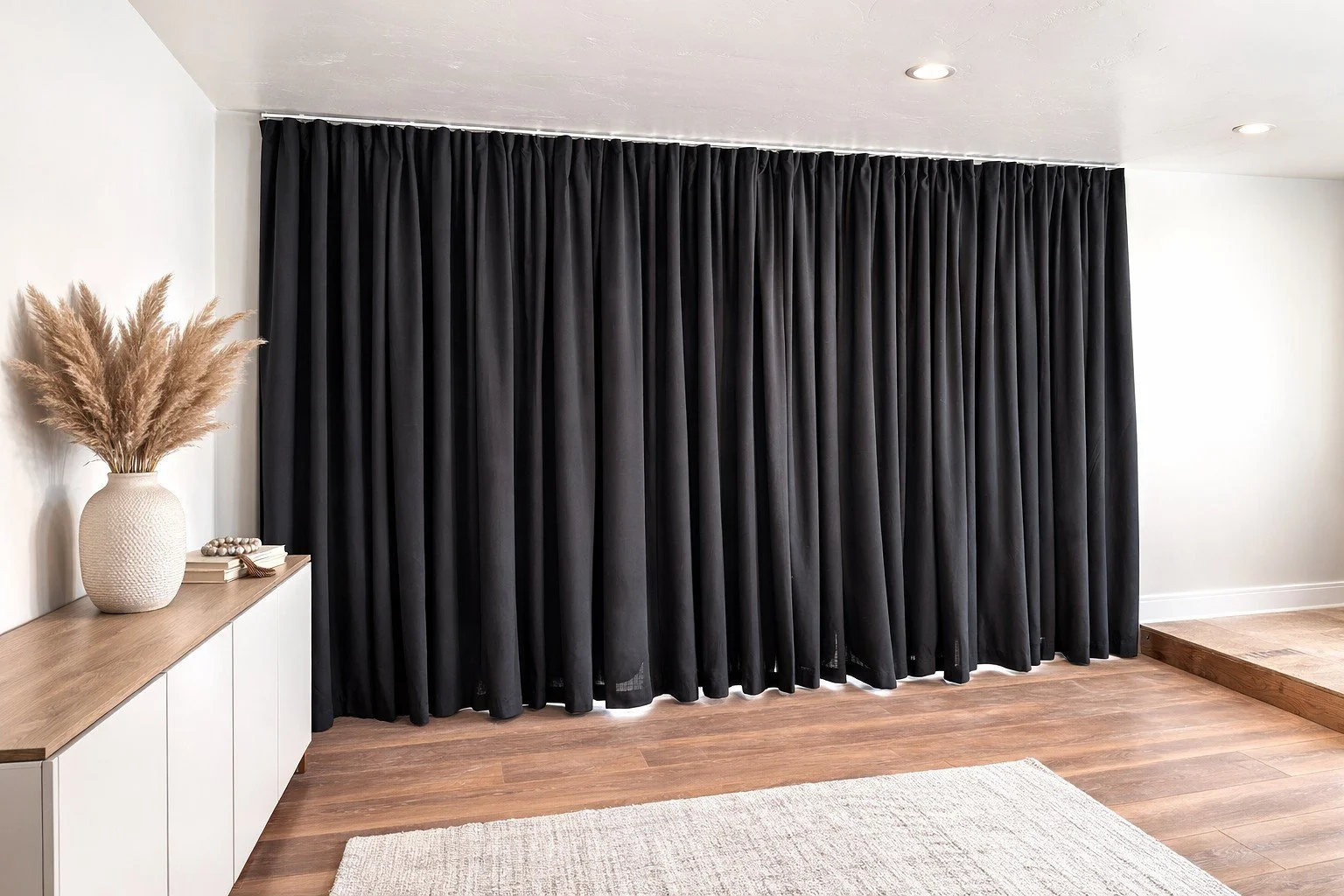 A room with black curtains hanging from ceiling to floor, a white sideboard with a vase of dried pampas grass and some decorative items, a light-colored area rug, and a hardwood floor, with recessed lighting in the ceiling.