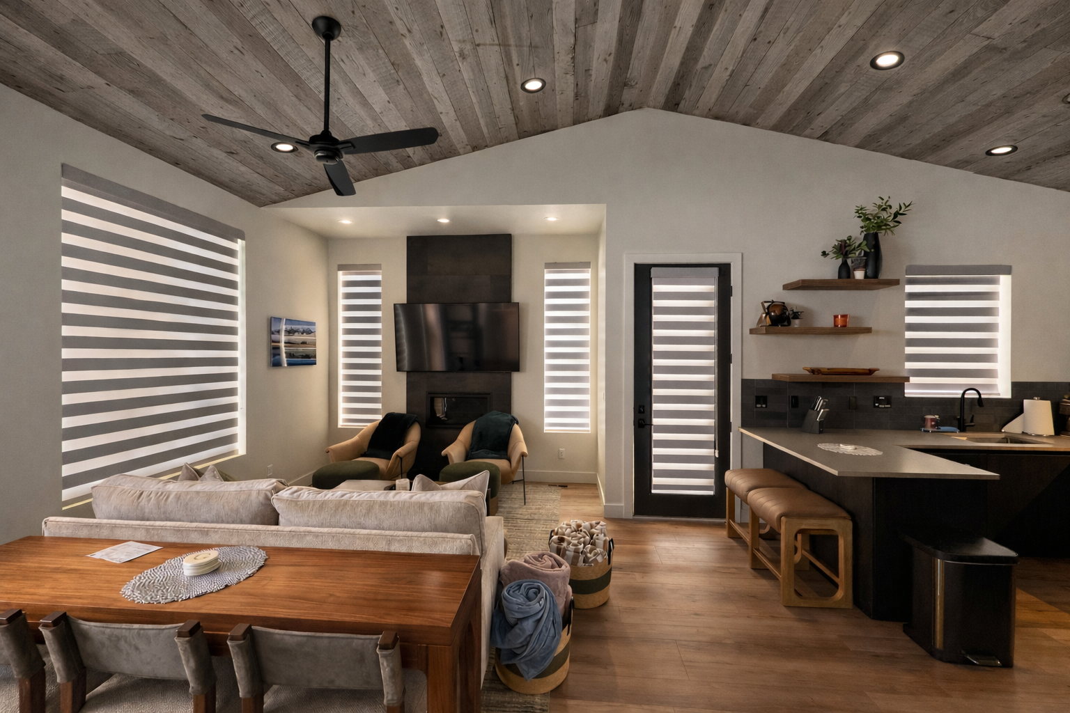 Open-concept living room with wooden ceiling, ceiling fan, large windows with striped blinds, and a kitchen area with a bar counter.