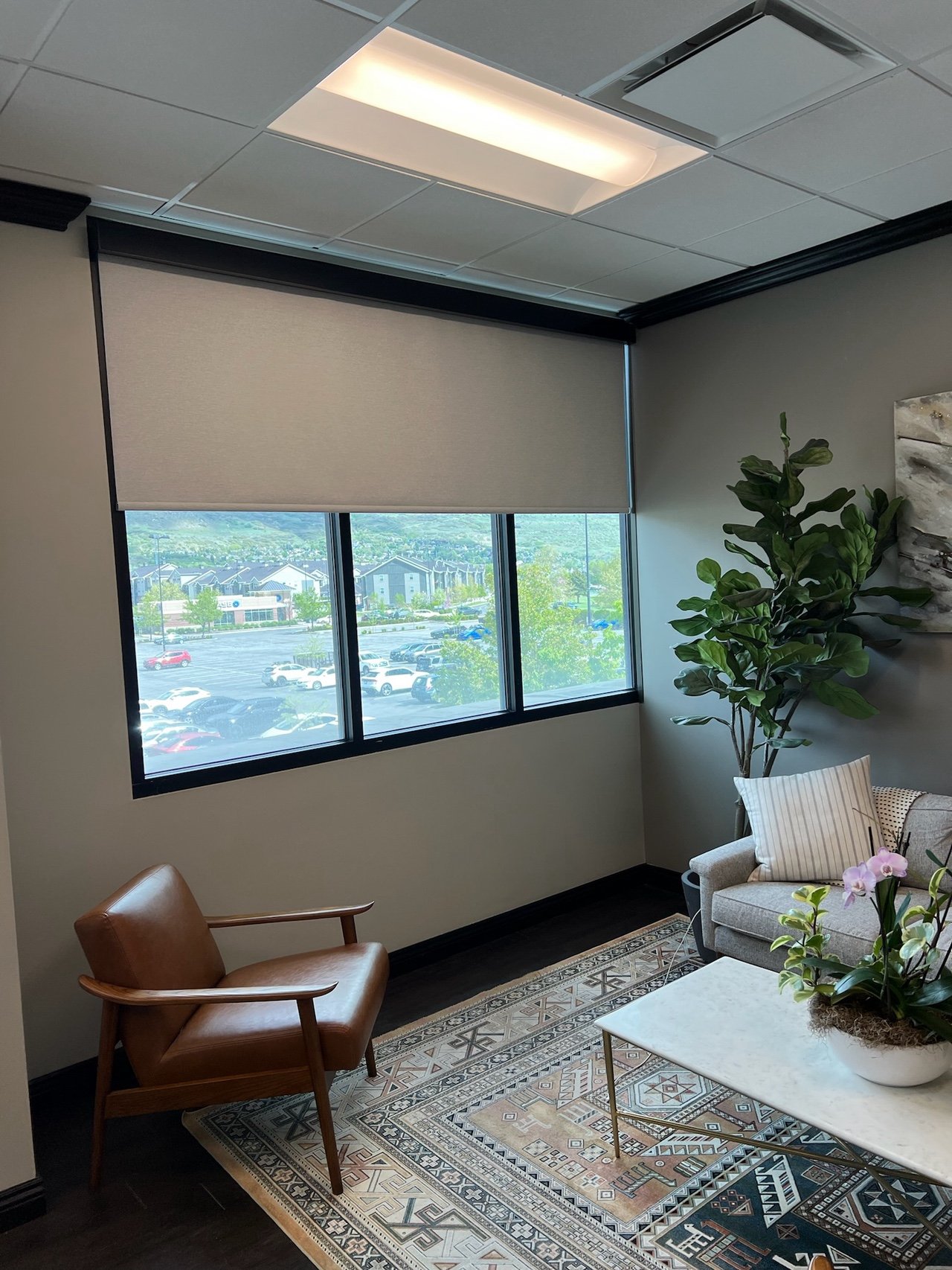 Interior of an office with a window, beige roller shade, potted plant, a brown leather armchair, gray sofa with striped pillow, a white coffee table with potted orchid, and patterned area rug.