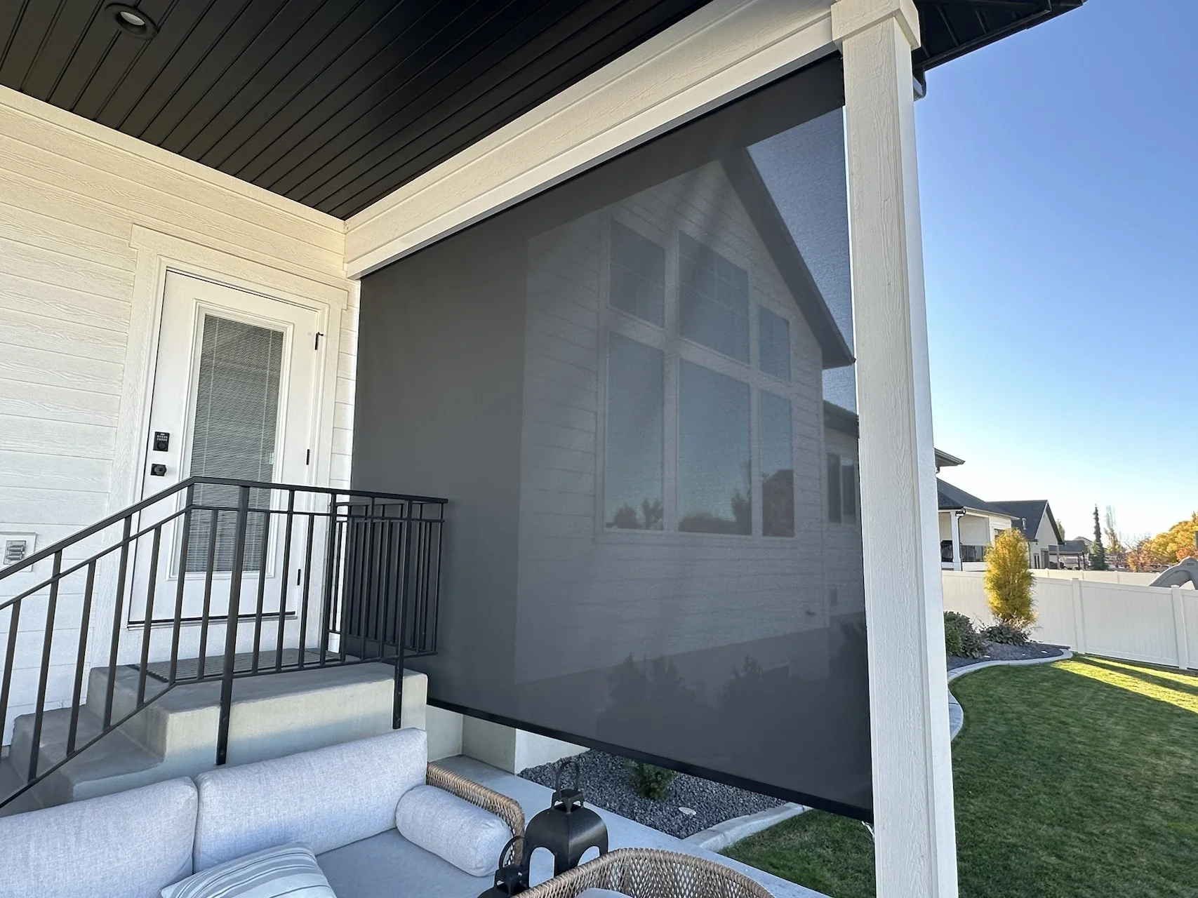 Outdoor patio with a black metal railing, a white door with a glass panel, and a large gray shade screen blocking part of the view of a house with white siding and large windows. There is a landscaped yard with grass, a small tree, and a white fence 