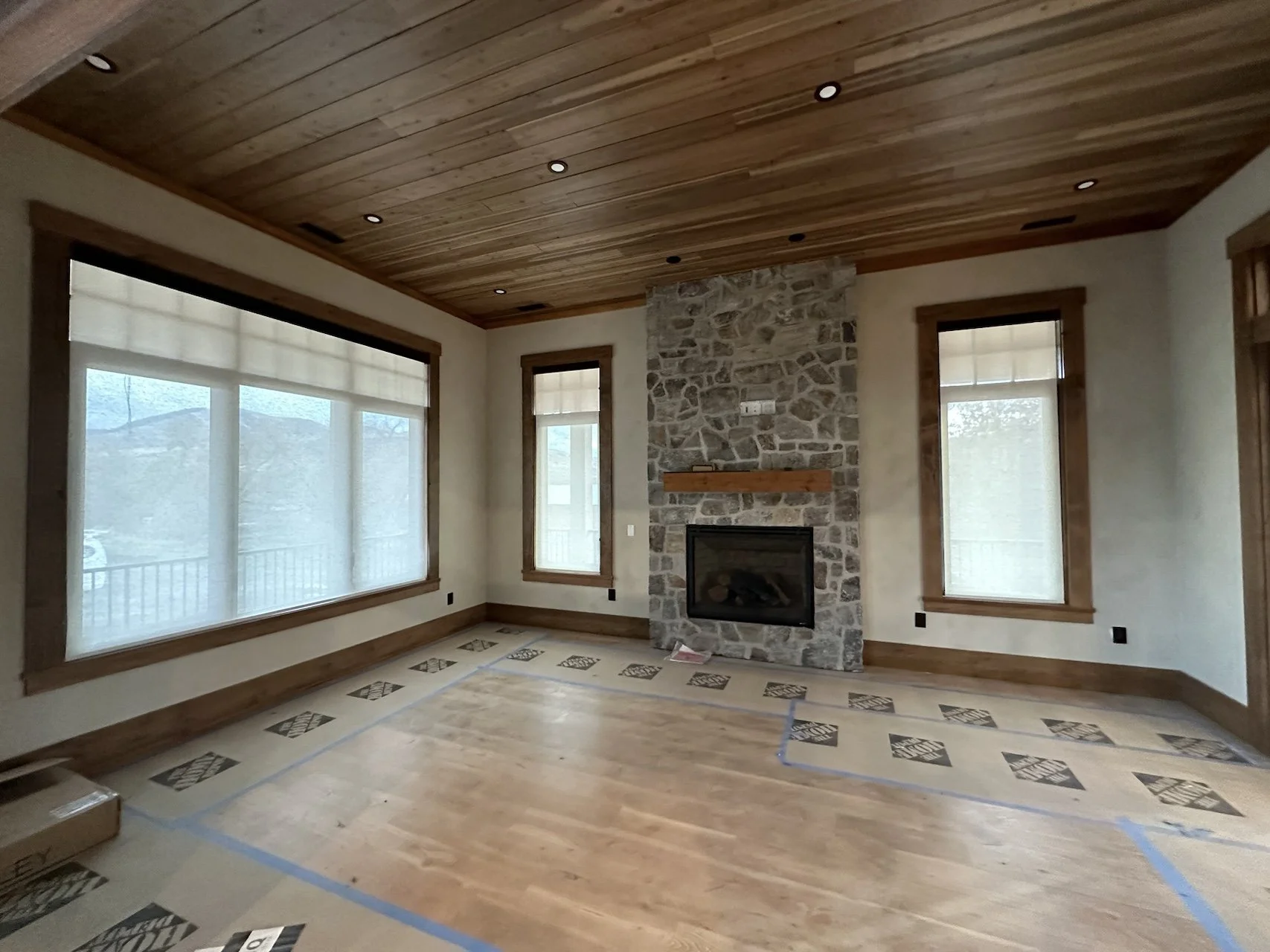 Living room with wooden ceiling, stone fireplace, large windows with shades, hardwood floor, and construction tape on the floor.
