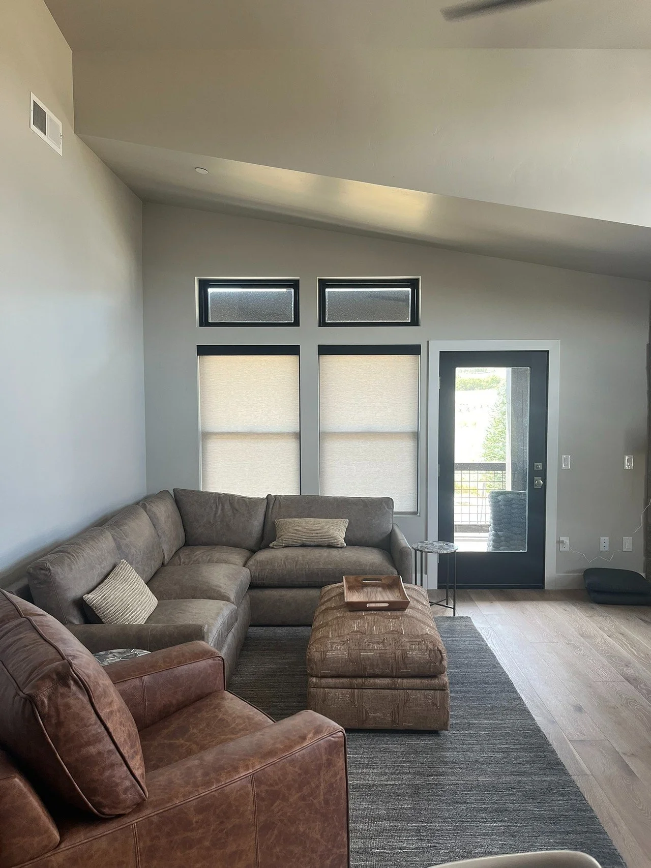 Living room with a sectional sofa, a leather armchair, an ottoman, a small side table, blinds on windows, and a door leading to an outdoor balcony.