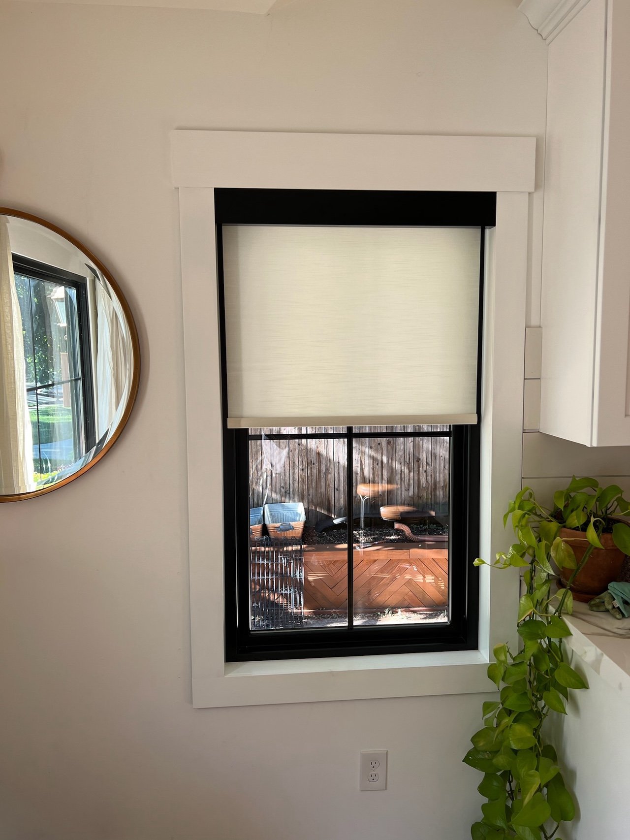 A window with a roller shade partially pulled down, revealing an outdoor patio with a wooden privacy fence, a small table, and two chairs. The window has a white frame with black trim, and there is a plant and a mirror on the wall nearby.