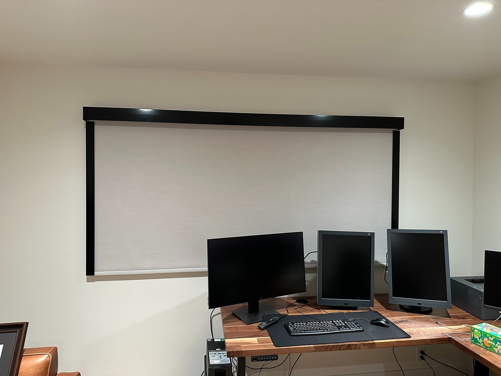 Office room with a large white projector screen, a wooden desk with three computer monitors, keyboard, mouse, and a tissue box.