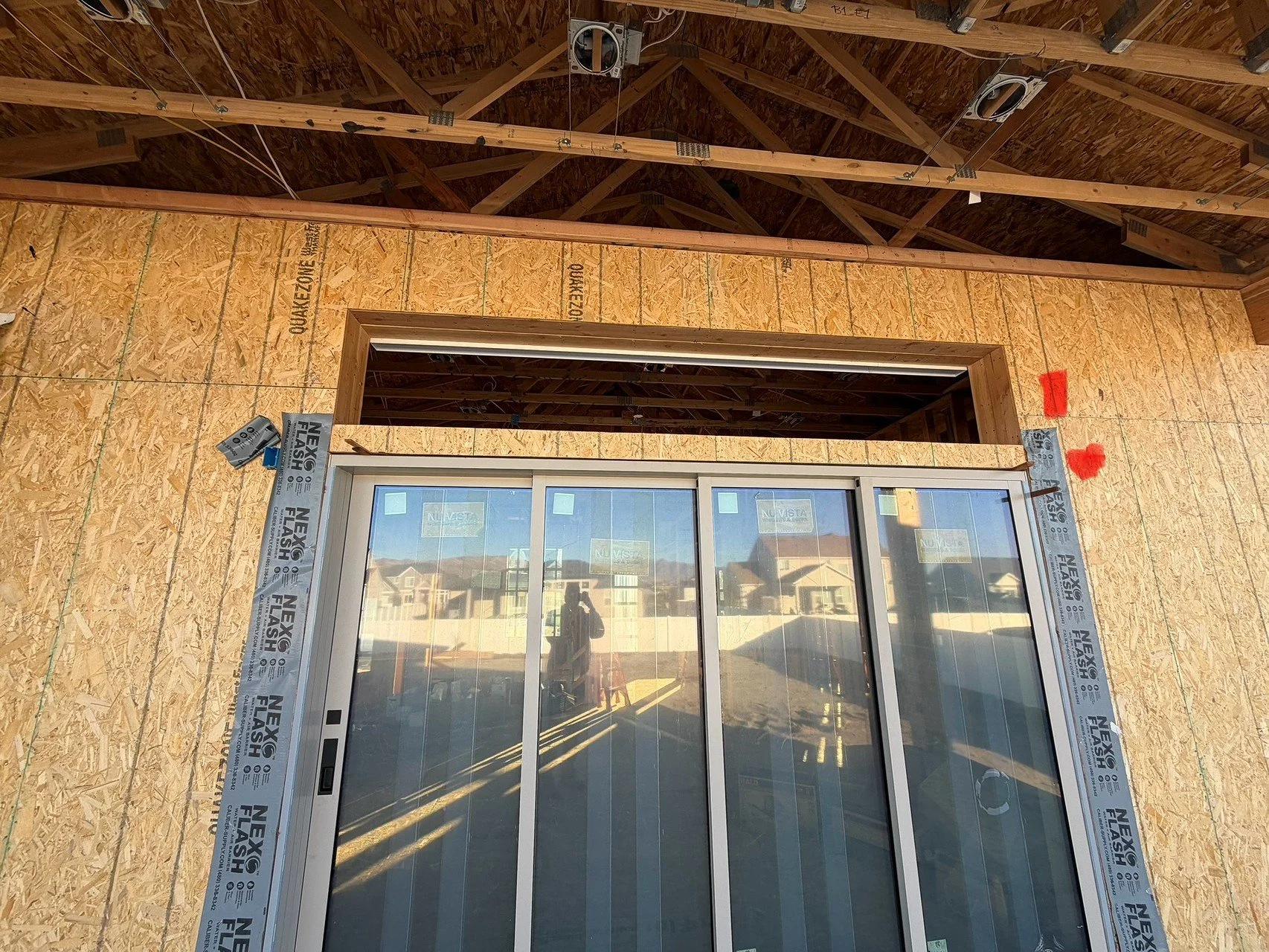 Construction site with a partially installed sliding glass door in an unfinished building. The wall is covered with oriented strand board (OSB) panels, and some red markings are visible. The ceiling is framed with wooden beams and trusses, and a refl