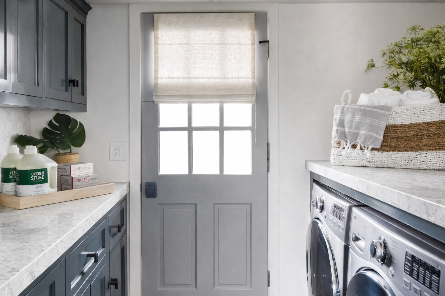 Laundry room with a door, washer and dryer, countertop with a basket, towels, and laundry supplies, and a window with a Roman shade.