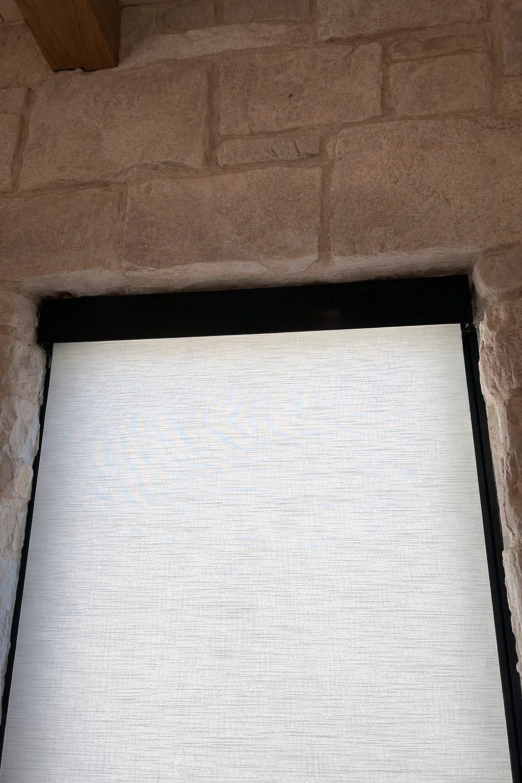 Close-up of a window with a stone wall and wooden beam above, partially covered by a frosted glass window shade.