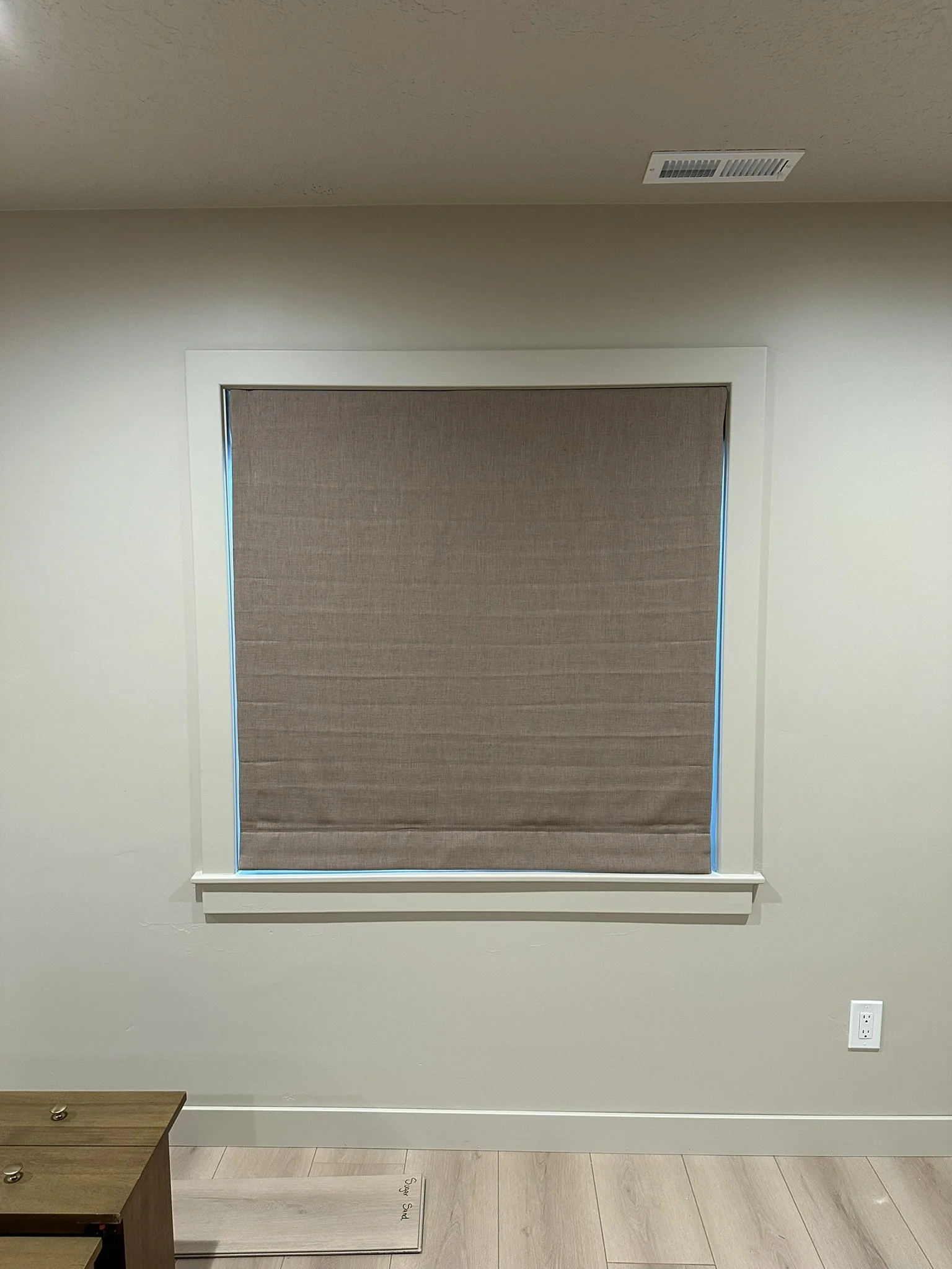 A wall with a window covered by a beige roller shade, and a wooden piece of furniture in the bottom left corner.