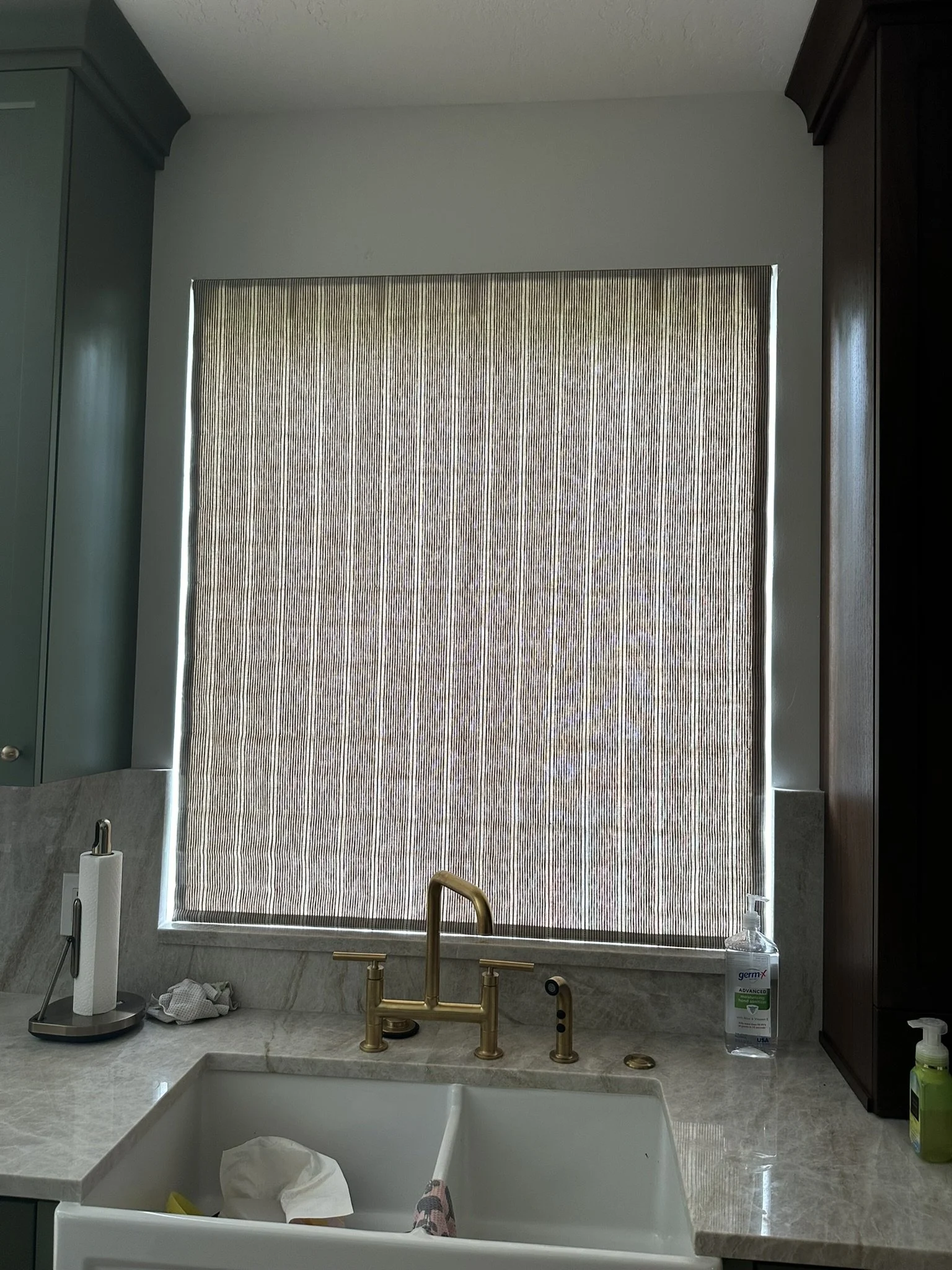 Kitchen window with closed beige vertical blinds, gold faucet and sink, soap dispenser, paper towel holder, and cleaning supplies on beige marble countertop.