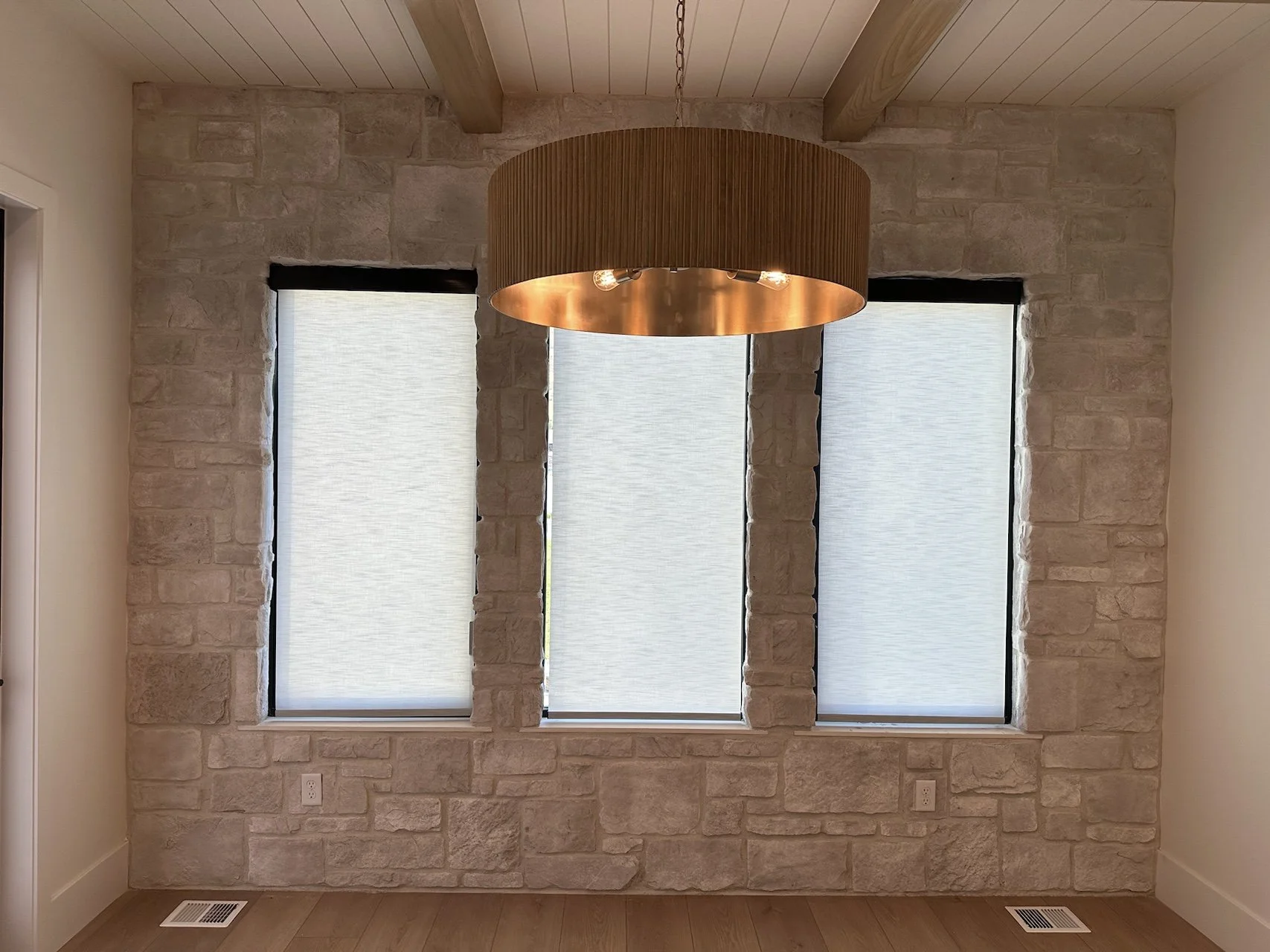 Interior view of a room with three large windows covered with translucent blinds, stone wall, wooden ceiling beams, and a large circular hanging copper light fixture.