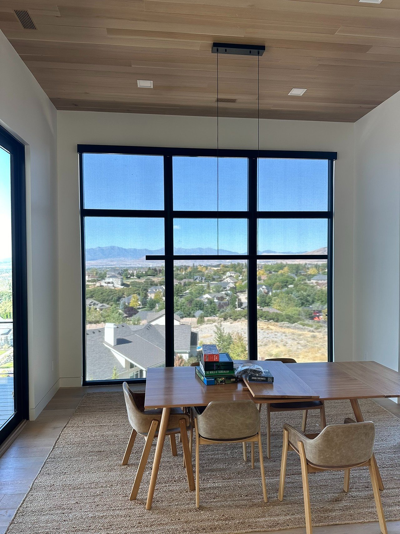 A dining room with a large window offering a view of a suburban neighborhood and distant mountains, a wooden table with chairs, and a modern pendant light hanging above the table.