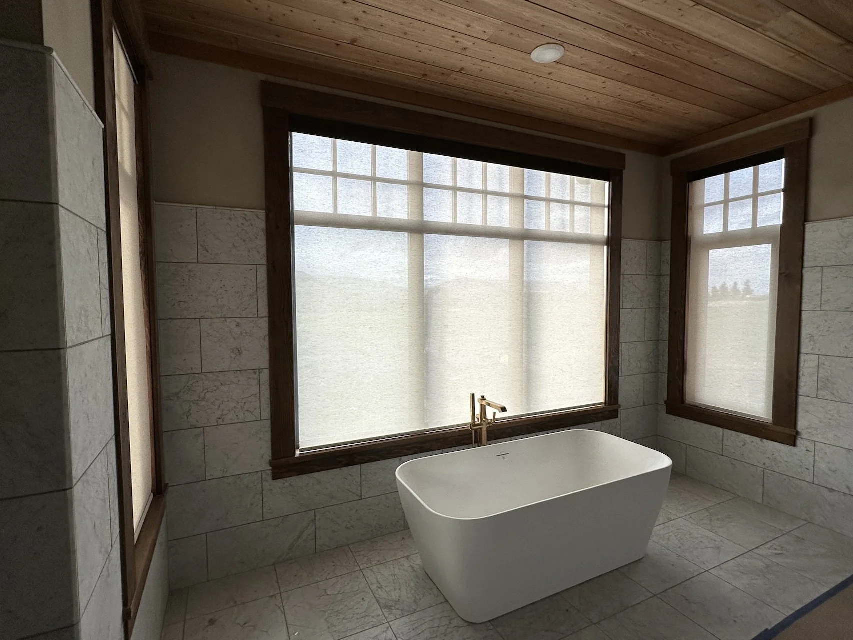 Modern bathroom with large window, freestanding bathtub, wood ceiling, and stone tile walls and floor.