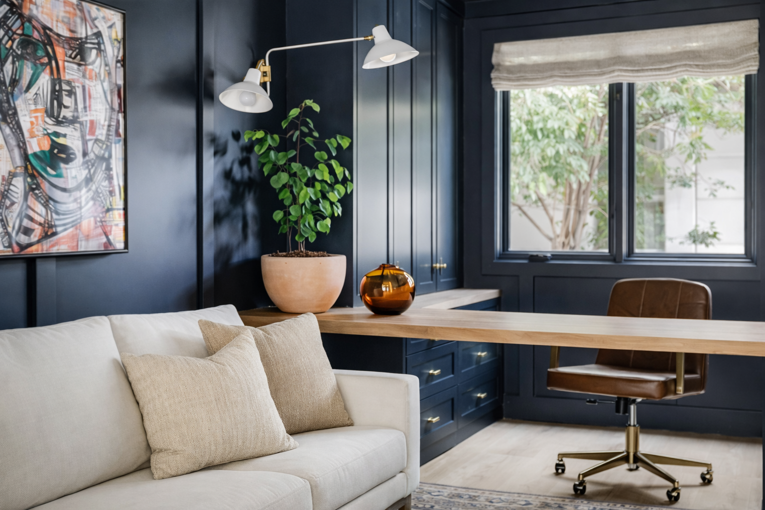 A modern home office with dark blue paneled walls, a beige sofa with matching pillows, a wooden desk, a brown swivel chair, a large window with greenery outside, a potted plant, a decorative amber vase, and colorful abstract artwork on the wall.