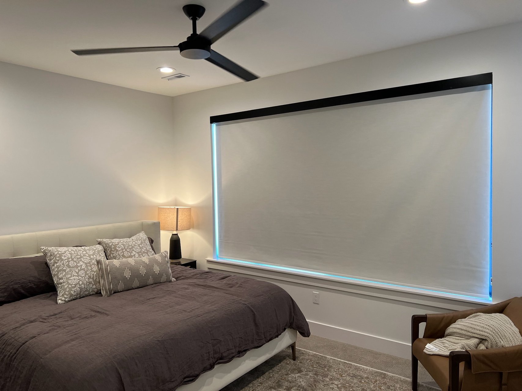 A bedroom with a large bed, dark bedding, and decorative pillows. A standing lamp is beside the bed. A window with a partially closed blind showing blue edges. A ceiling fan and recessed lighting are on the ceiling, and a beige armchair with a blanke