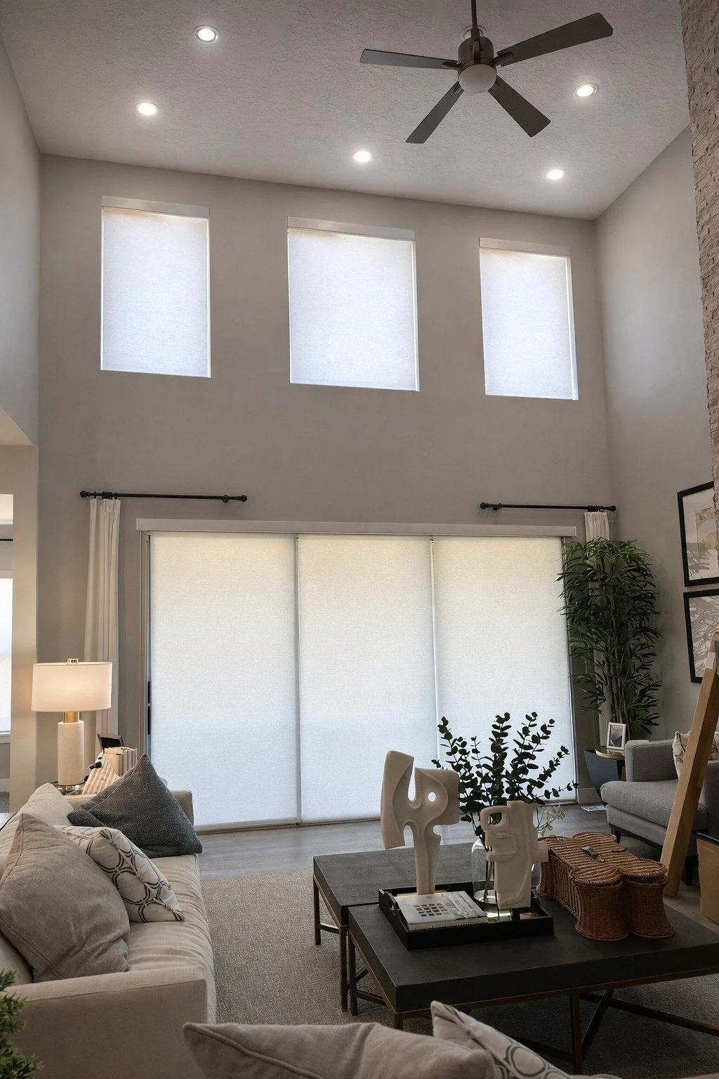 Living room with high ceiling, three small windows near the ceiling, a ceiling fan, large sliding glass door with white roller shades, a gray sofa with patterned and solid pillows, a side table with a lamp, abstract sculptures on a coffee table, hous