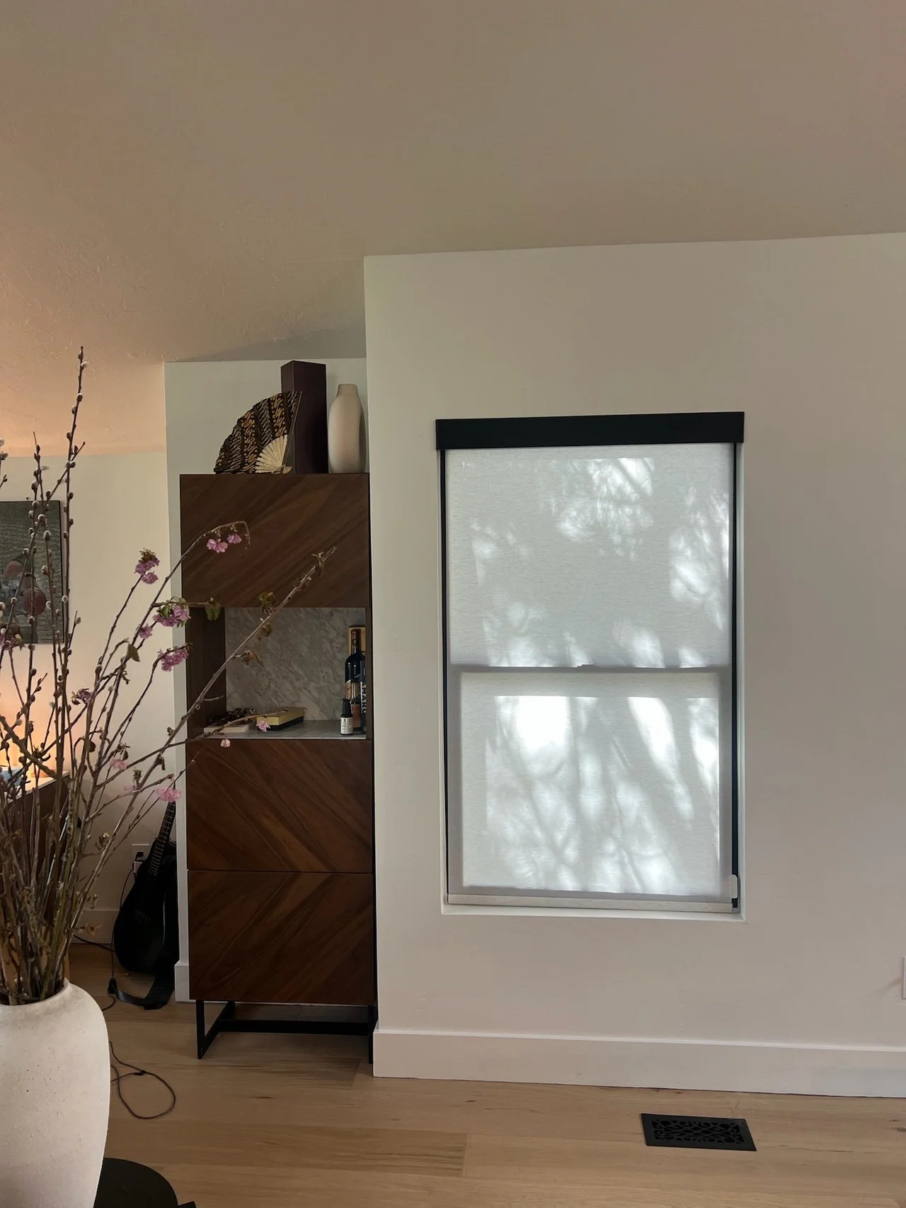 A section of a living room with a window covered by a light-colored roller shade, partially showing sunlight and tree shadows outside. To the left, a tall wooden bookshelf with decorative items and books. A large white pot with pink flowering branche