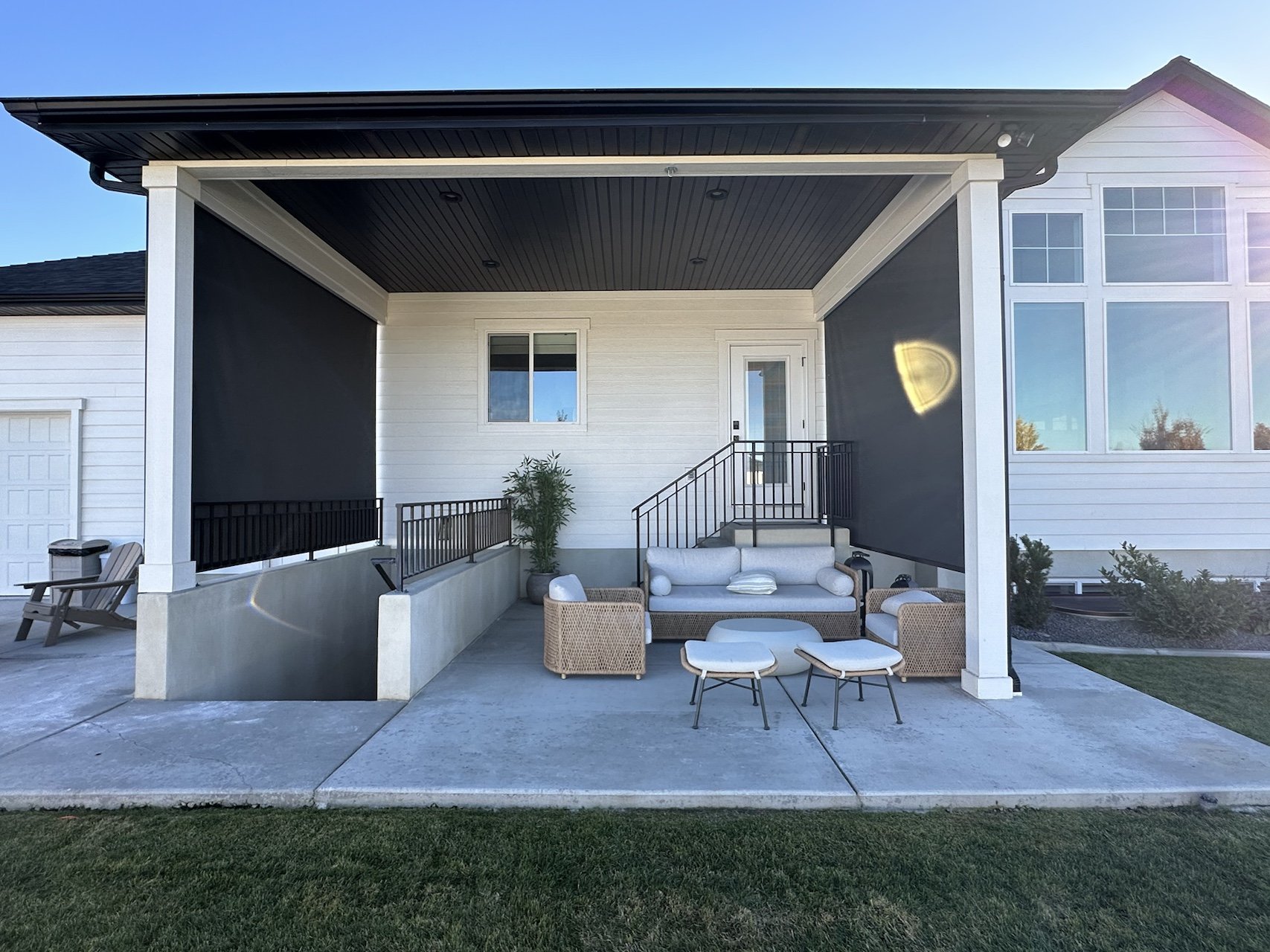 Front porch of a modern house with outdoor seating, including a sofa and small tables, under a large covered area with black ceiling panels, white walls, and large windows.