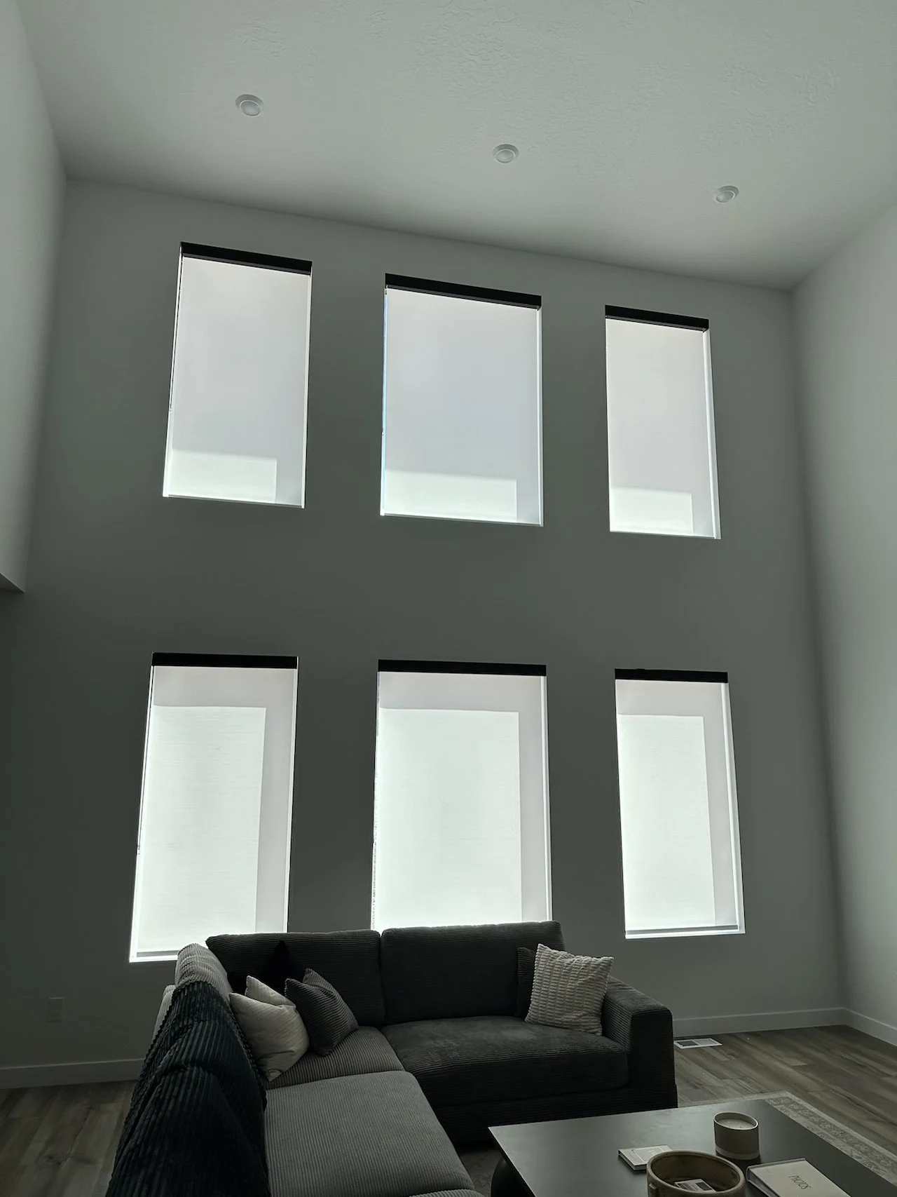 Living room with six tall windows with blackout blinds, white walls, and wooden flooring. A sofa with several pillows and a coffee table are visible.