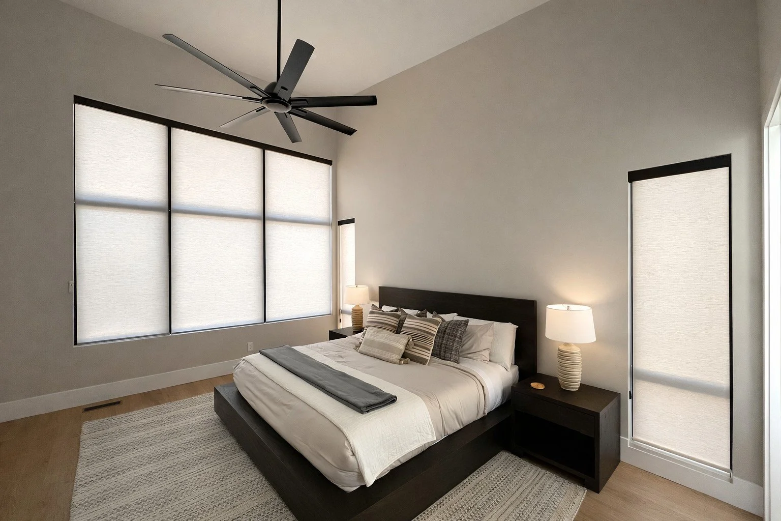 Modern bedroom with large window blinds, dark wood bedframe, white bedding, striped pillows, matching nightstand, table lamp, ceiling fan, and light-colored rug.