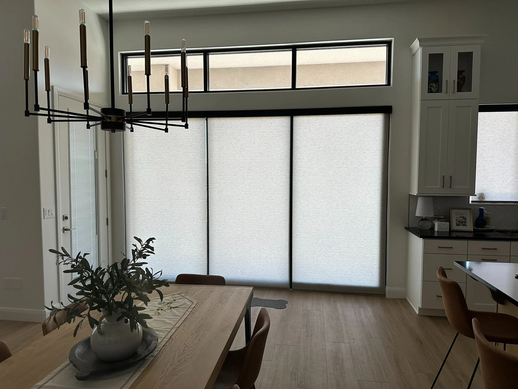 Dining area with a wooden table, a vase with greenery, and a black chandelier, adjacent to a kitchen with white cabinets and a large frosted glass sliding door cover.