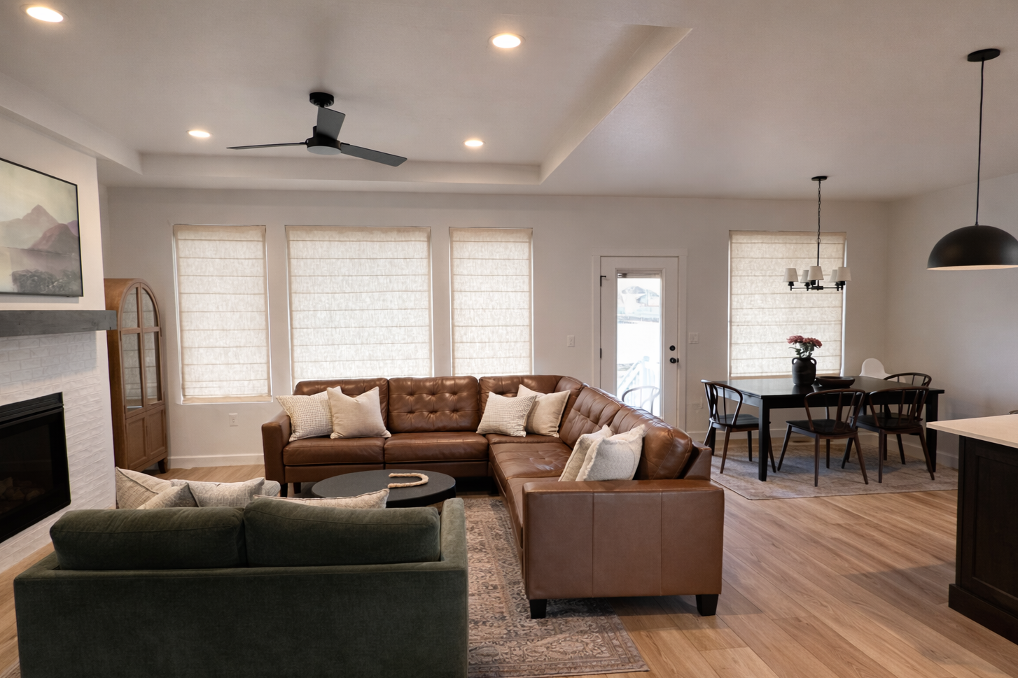 Living room with a brown leather sectional sofa, a green armchair, a black coffee table, and a fireplace. There are three large windows with beige shades, a ceiling fan, and a dining area with a black table, six chairs, and a hanging light fixture.