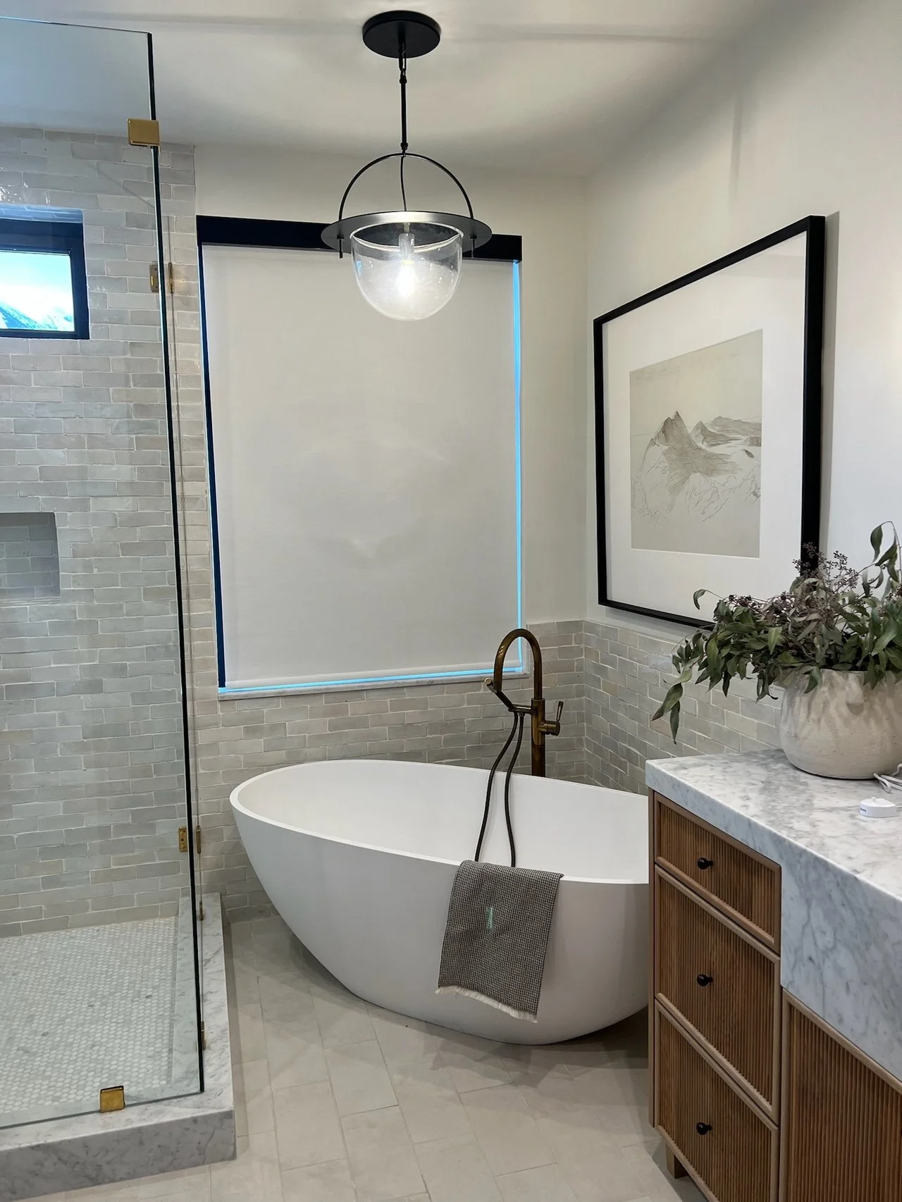 Modern bathroom with a freestanding bathtub, a glass shower enclosure, a wooden vanity with a marble countertop, a large framed mountain artwork on the wall, and a potted plant.