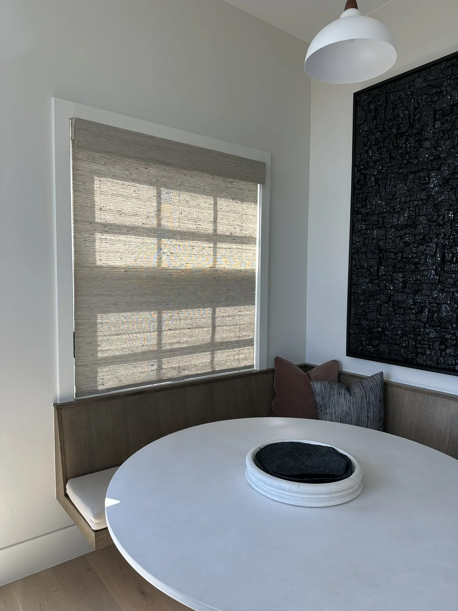 Indoor corner with a round white table, a brown cushioned bench with pillows, a window with beige blinds casting shadows, a white ceiling light, and a large black textured art piece on the wall.