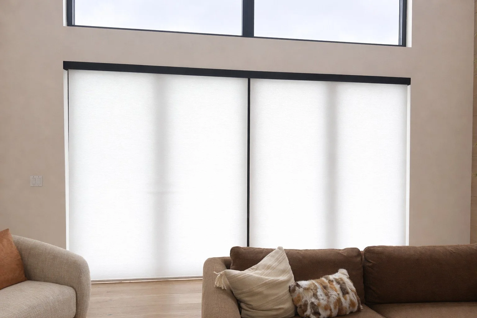 Interior living room with large window covered by white blinds, beige walls, and brown sectional sofa with pillows.
