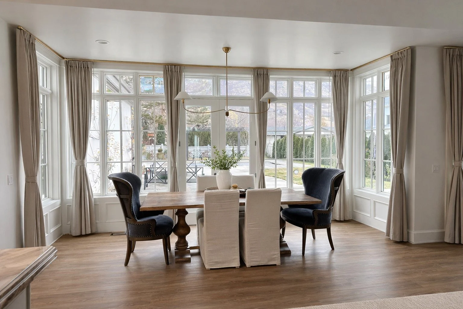 Dining room with large windows, light-colored curtains, a wooden table, six chairs with some covered in white slipcovers, a white vase with greenery on the table, and a modern chandelier.